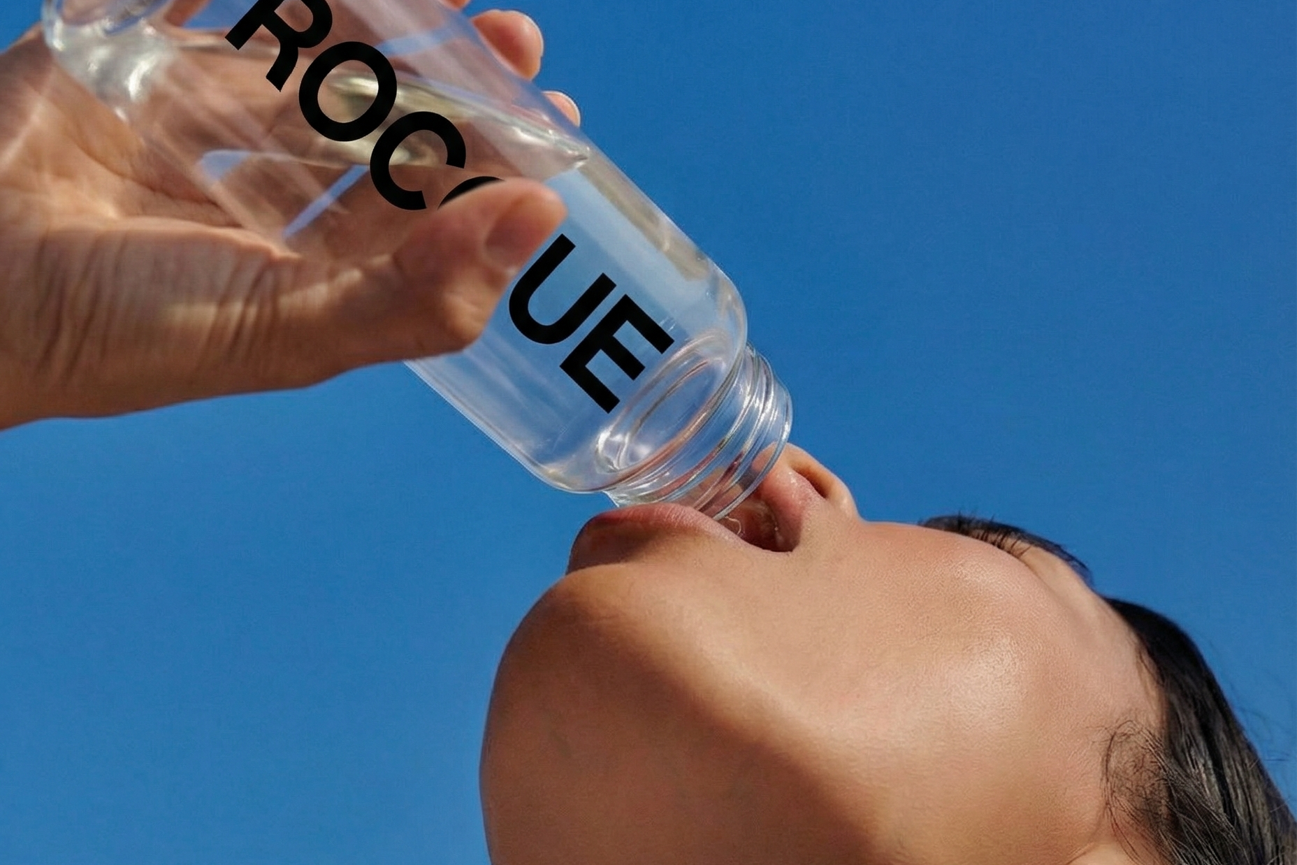 Person drinking from a clear water bottle with 'ROCQUE' branding against a blue sky.