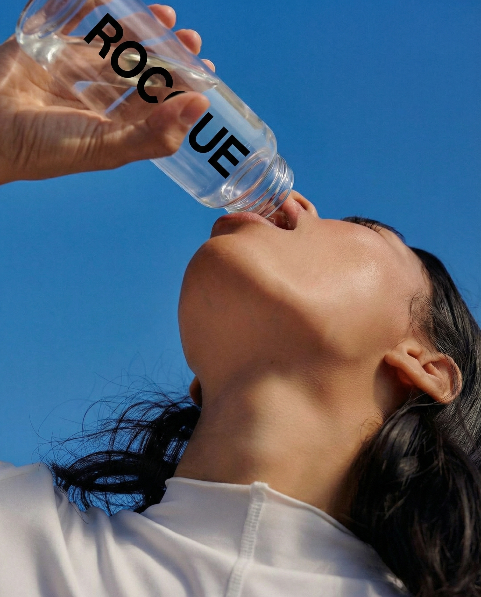 Person drinking water from a bottle labeled 'ROCQUE' against a clear blue sky