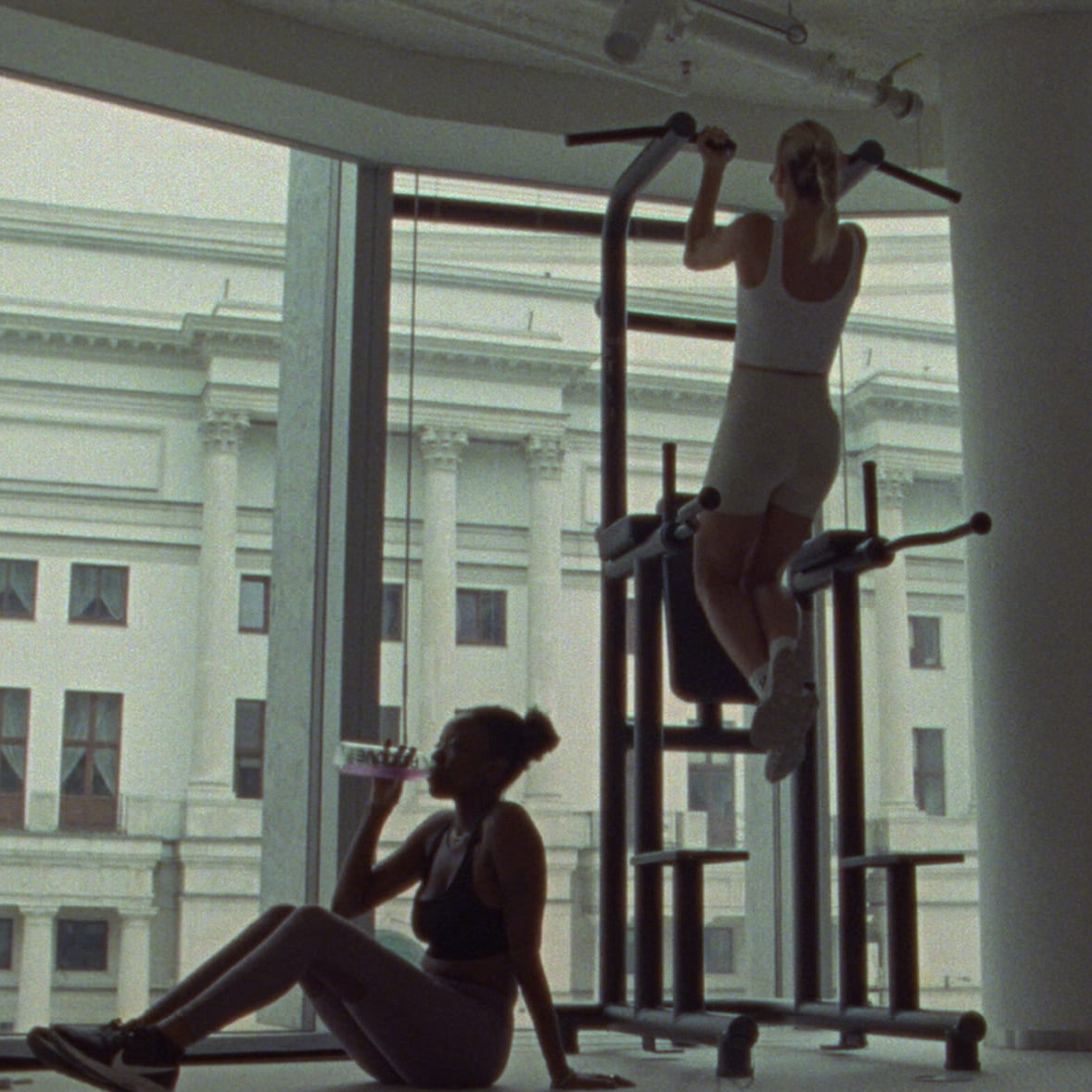Person exercising on a pull-up bar in a gym with large windows.